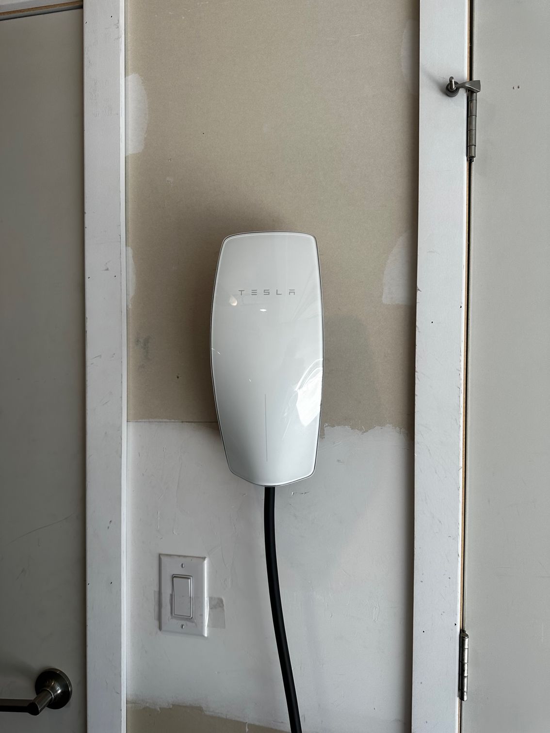 White wall-mounted device with a black cable beside a light switch and door frame