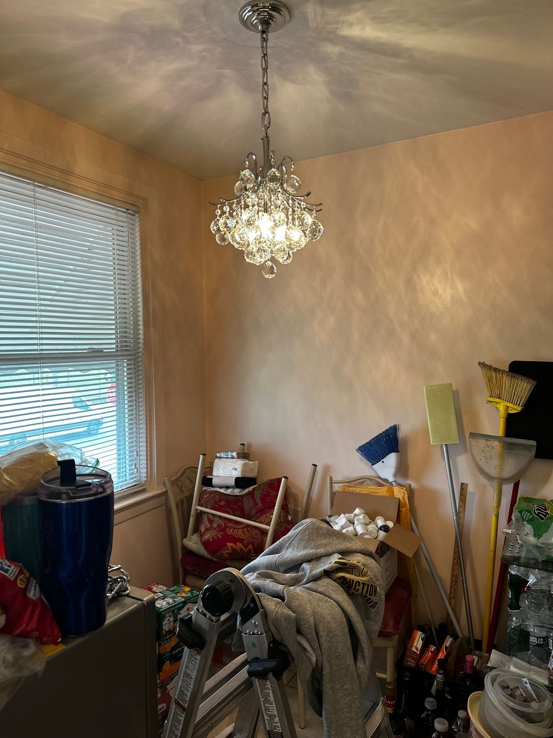 Cluttered room with chandelier, window blinds, and stacked household items against beige walls.