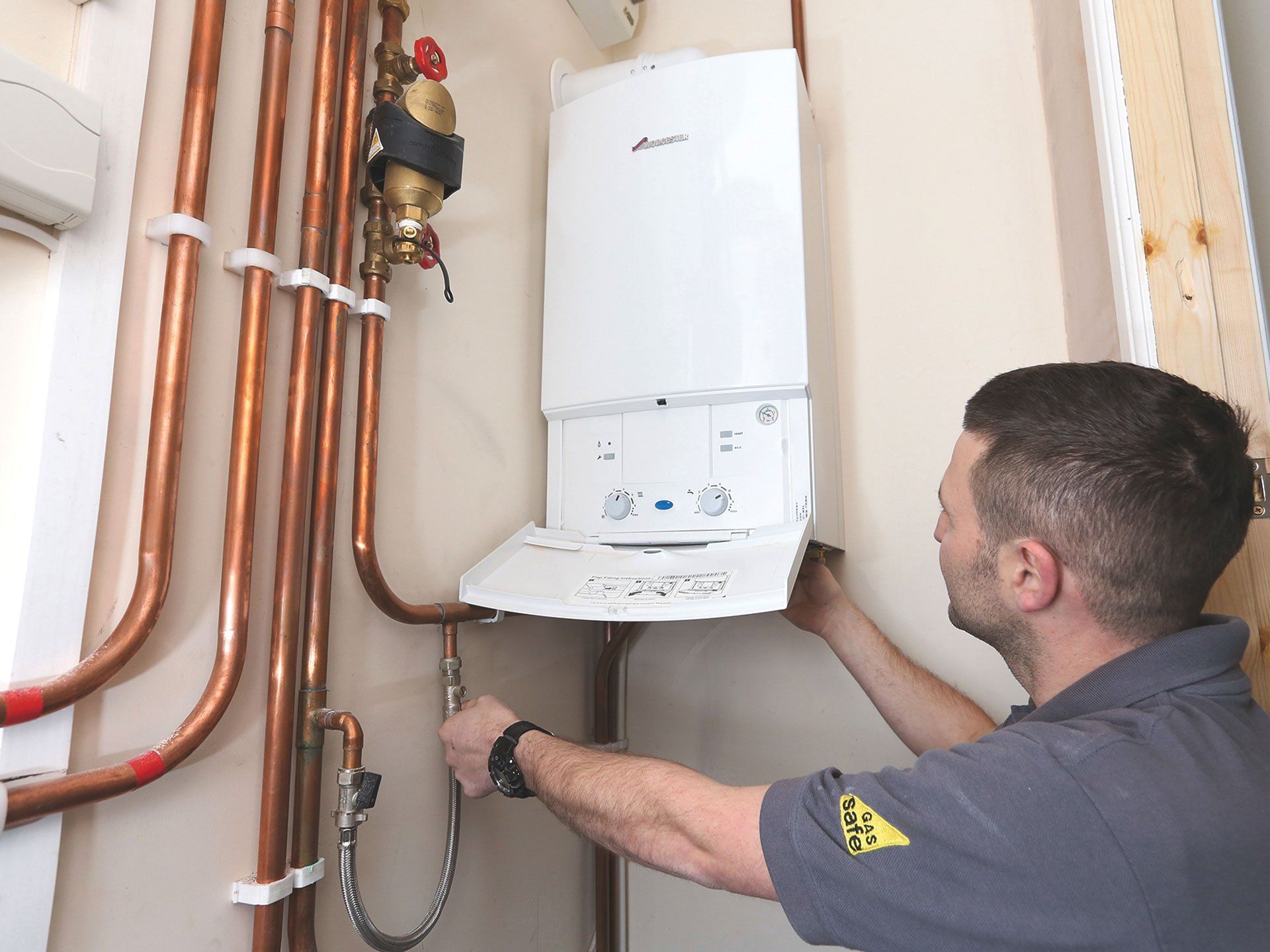 Technician installing a white wall-mounted boiler beside copper pipes in a utility room