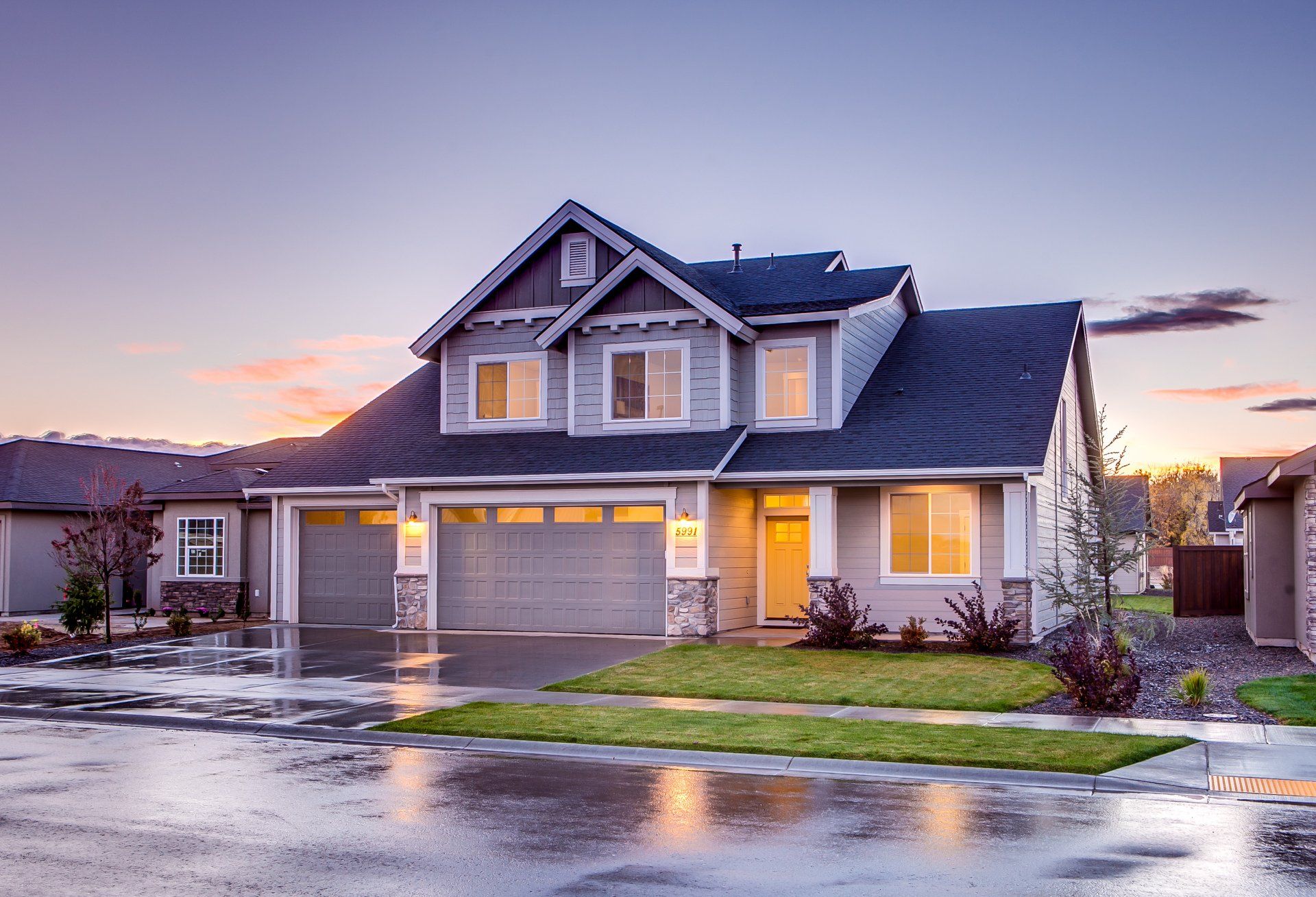 Suburban two-story house with lit windows, garage, and wet driveway at sunset