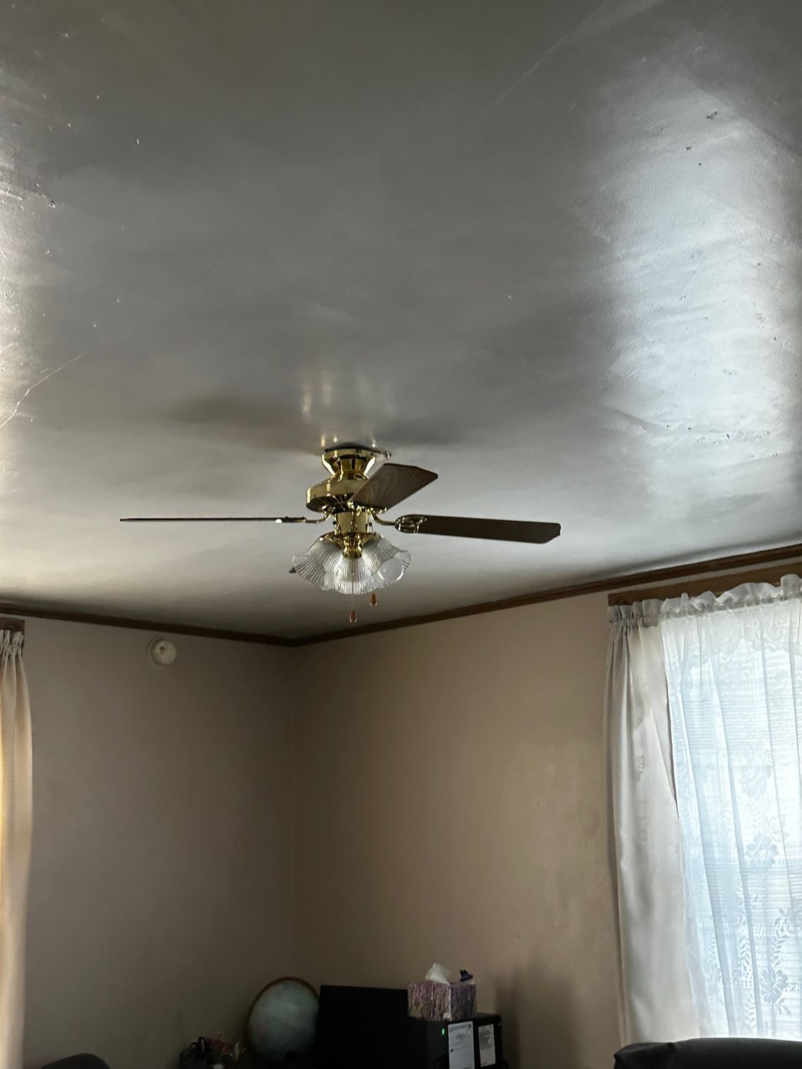 Living room ceiling with brass ceiling fan, beige walls, and a sunlit window with curtains