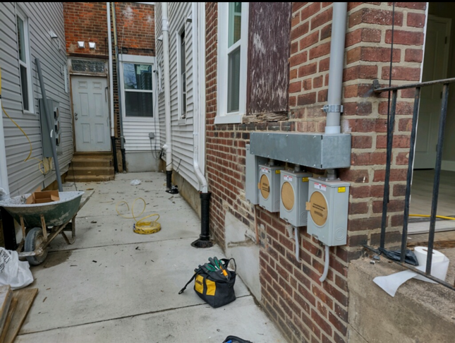 Narrow concrete alley between houses with utility meters on a brick wall and a wheelbarrow in the foreground