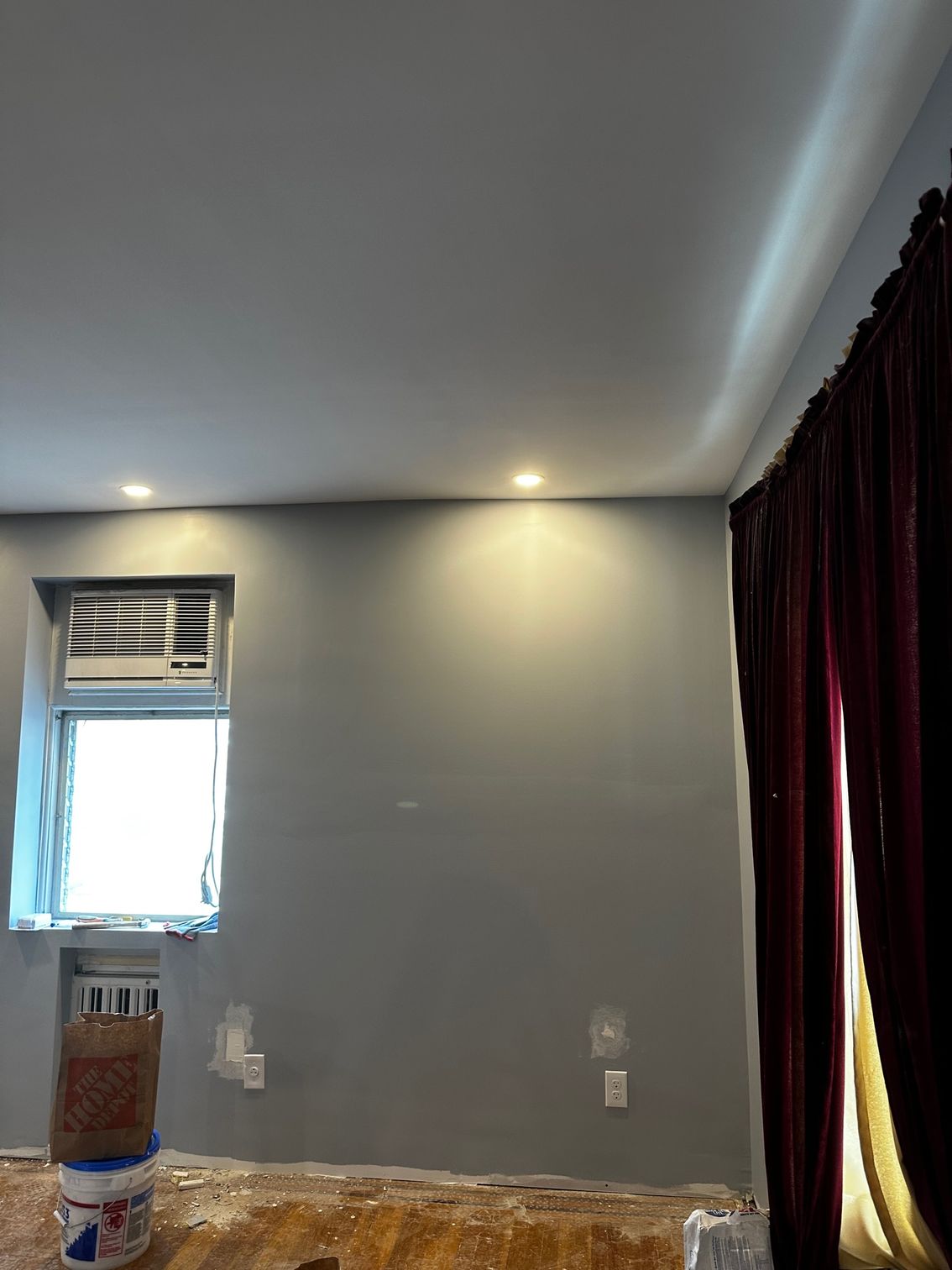 Empty room with gray walls, recessed ceiling light, window, and dark curtains; floor partly covered during renovation
