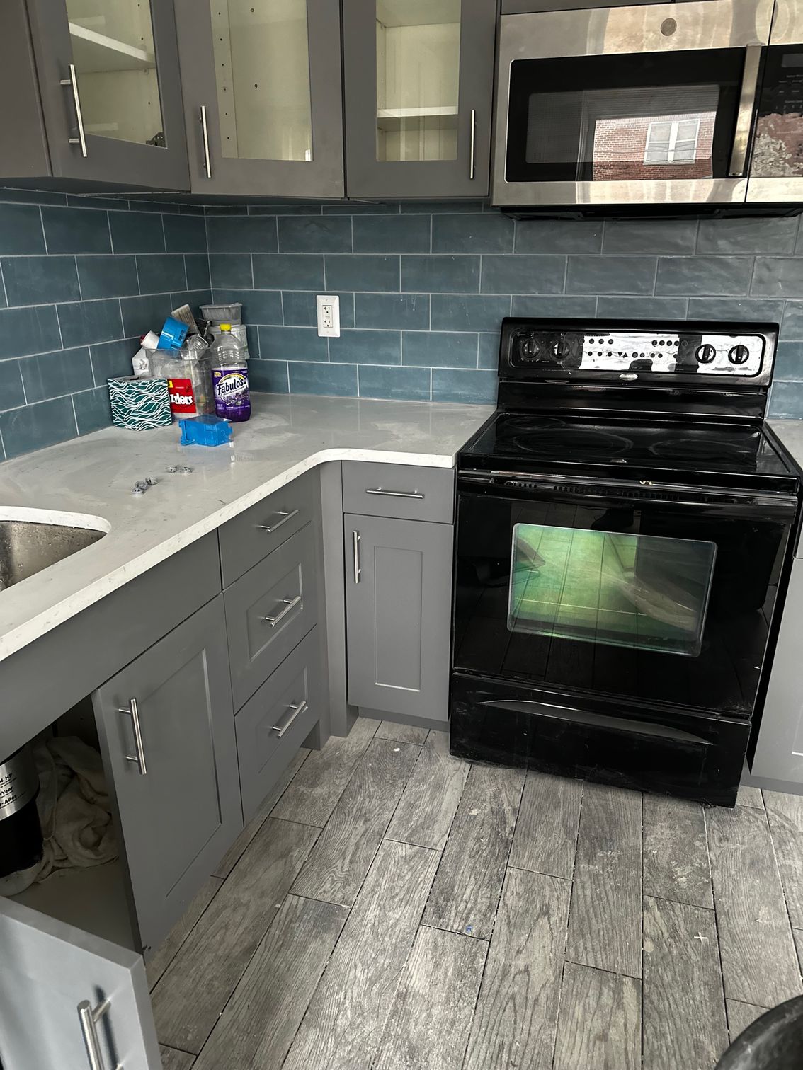 Small kitchen with gray cabinets, white counters, black stove and microwave, and blue tile backsplash.