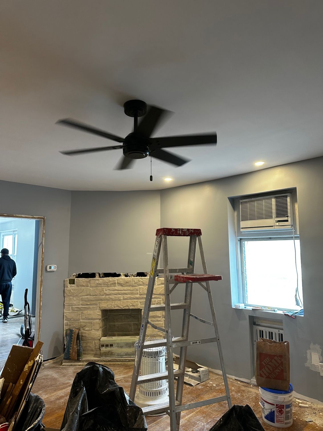Room under renovation with ladder, exposed fireplace, ceiling fan, and window with blinds