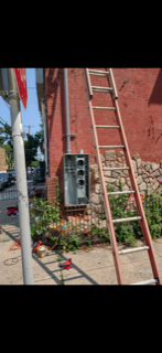 Red brick building wall with a leaning ladder, gray utility box, and flowers by the base