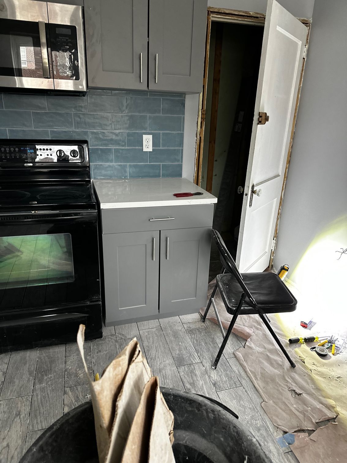 Small kitchen with gray cabinets, black stove, tiled backsplash, and open doorway; debris on floor.