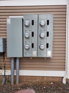 Two gray electric utility meter boxes mounted on an exterior wall with tan siding and conduit pipes below.