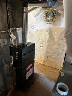 Basement furnace beside a white wall with exposed ductwork and coiled cable.