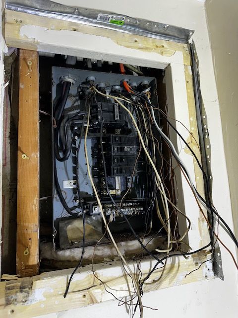 Exposed electrical panel with tangled wires inside a wall opening, surrounded by torn drywall and trim