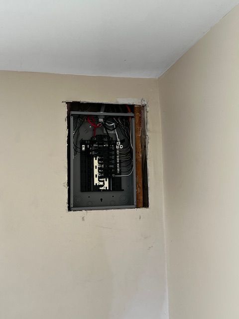 Open electrical breaker panel recessed in a beige wall corner with exposed wiring.