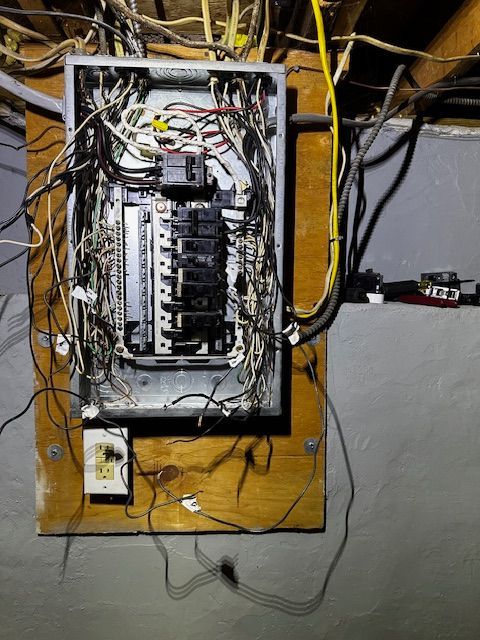 Open electrical breaker panel with many tangled wires mounted on a wooden board.