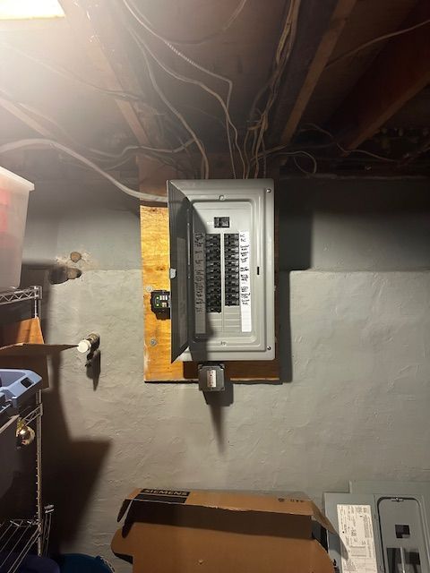 Electrical breaker panel mounted on a basement wall with exposed wiring above and storage boxes nearby