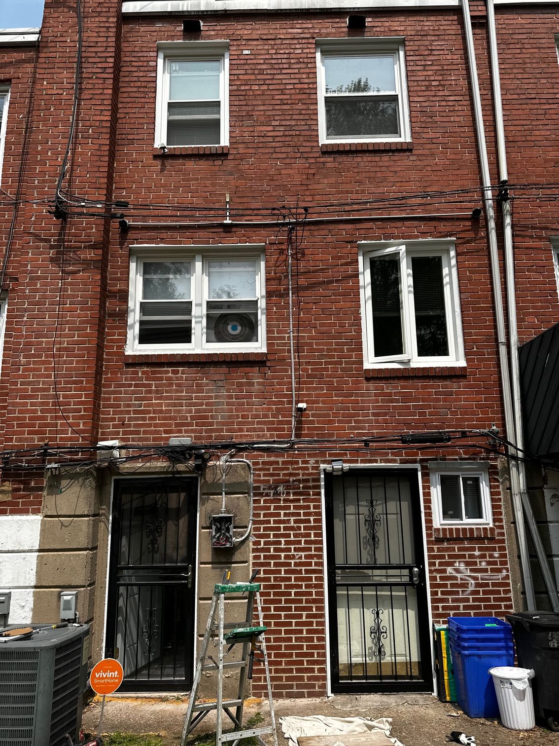 Old red-brick rowhouse facade with boarded lower windows, a metal door, and trash bins out front