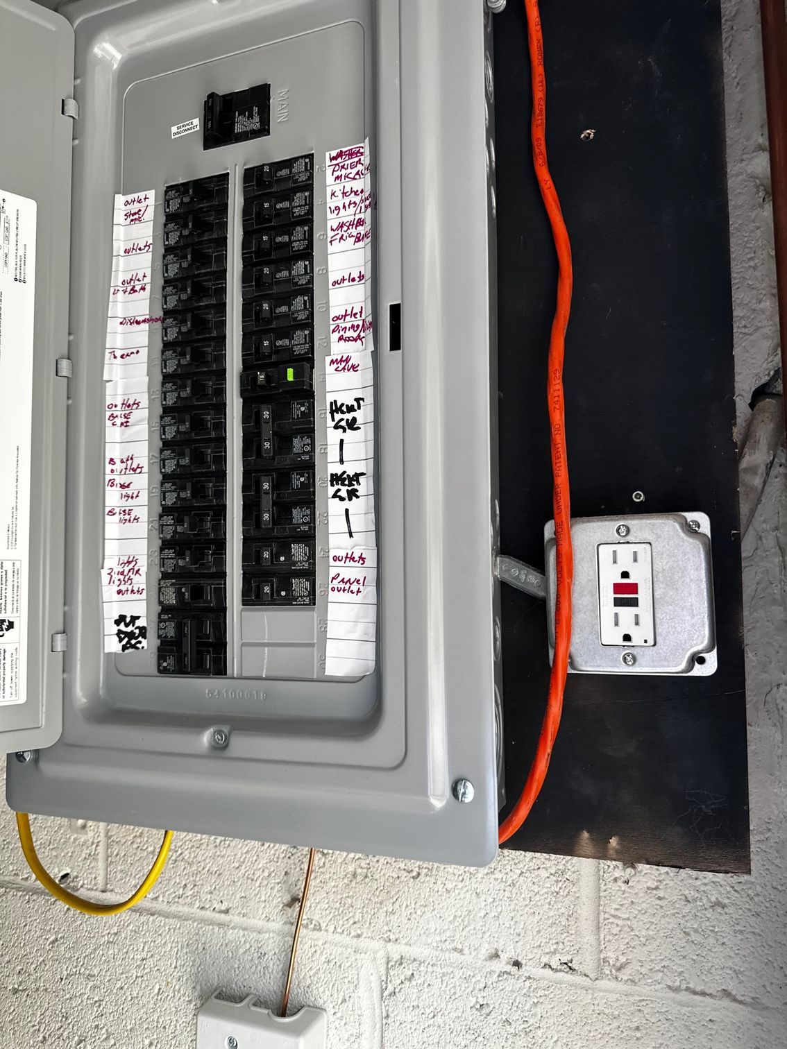 Open electrical breaker panel with labeled circuits and nearby outlet box on a concrete wall.