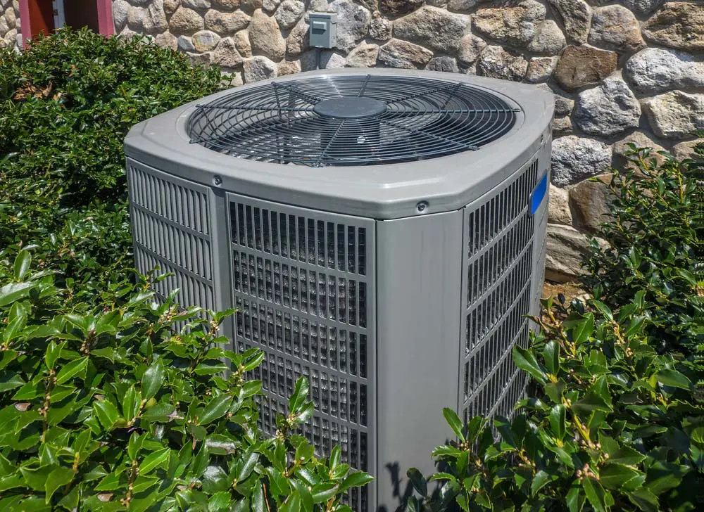 Outdoor air conditioner unit beside a stone wall, partially surrounded by green shrubs.
